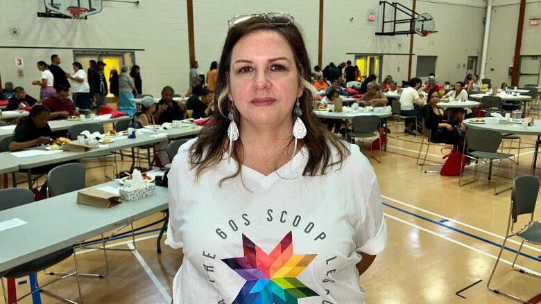 A woman looks into the camera and smiles. She wears a white shirt that reads 