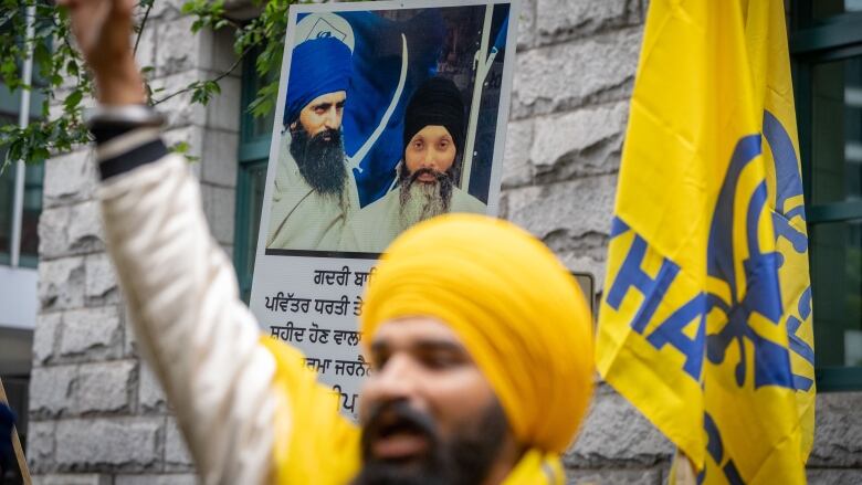 A man is pictured with his fist raised during a protest, behind him a portrait of Shaheed Bhai Hardeep Singh Nijjar.