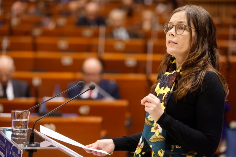 A woman wearing glasses makes a speech at a podium.