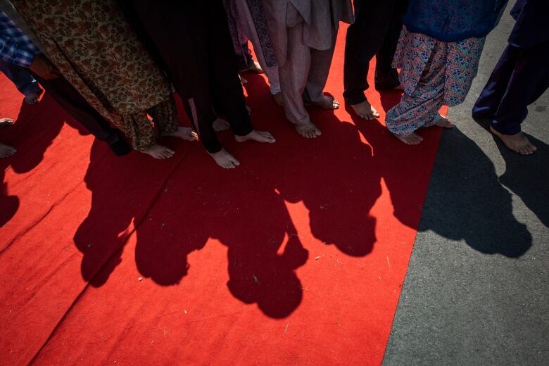 A lineup  of barefoot women at a Sikh funeral cast their shadows towards the camera on a red carpet.