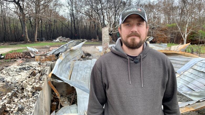 A man in a ball cap stands in front of a burned out house. 