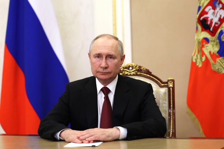 A man in a suit and tie sits in a chair, looking glum, his hands folded in front of him on a piece of paper on a table. He is flanked by two flags in Russian colours.