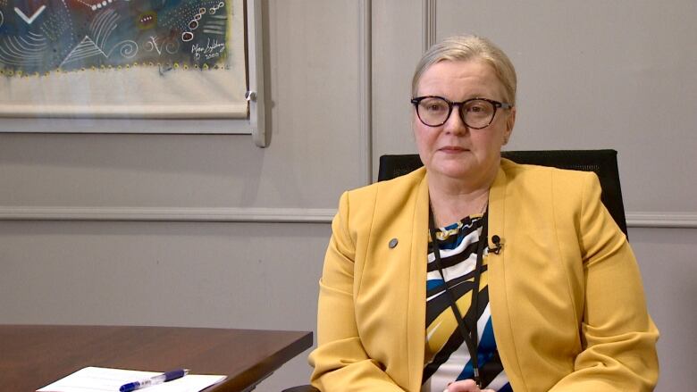 A woman in a pale yellow jacket sits next to a desk.