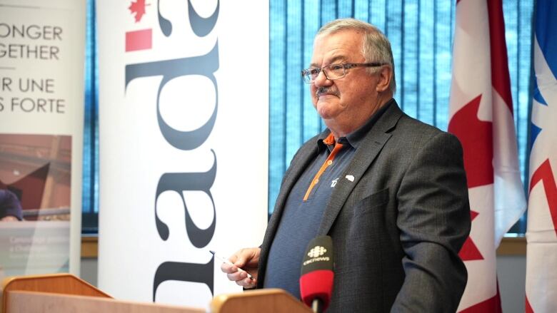 A man in a black blazer and blue shirt stands in front of a podium 