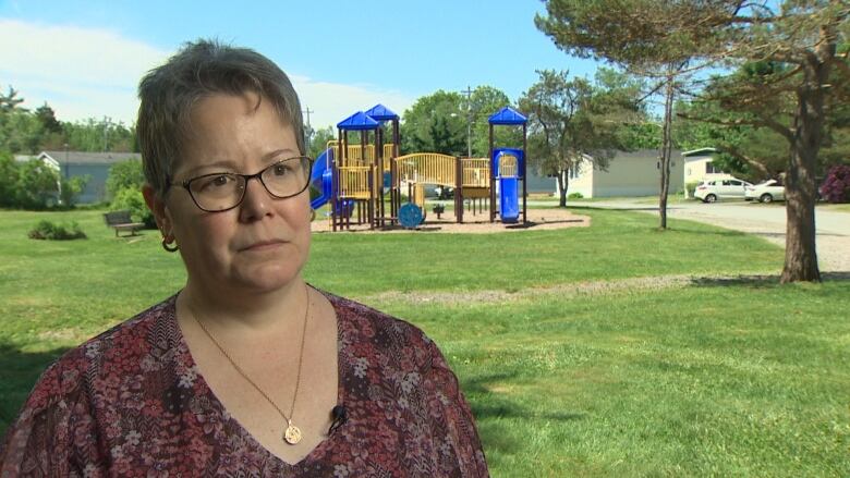 A white woman with short grey hair and glasses wears a red top and gold necklace. Behind her can be seen a green field, playground and mobile homes.