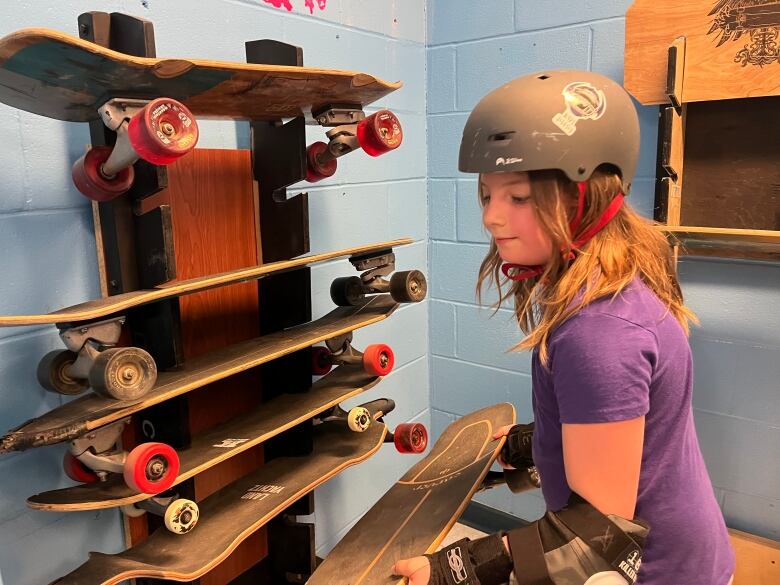 A girl in a helmet picks one skateboard from a rack of skateboards.