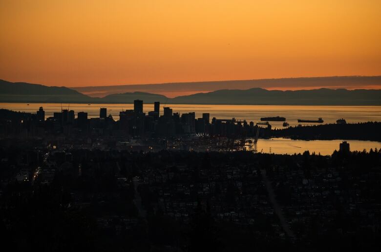 A dark orange sky is seen above a nighttime silhouette of downtown Vancouver and Burrard Inlet.