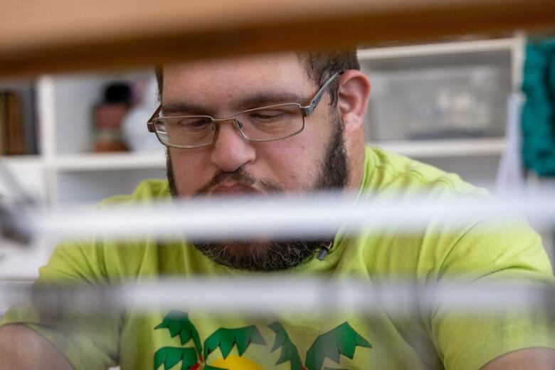 A closeup shot shows a bearded person with a yellow shirt focusing on weaving. 