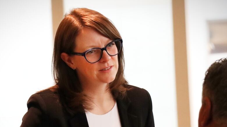 A woman with glasses and a brown sport jacket speaks with someone else.