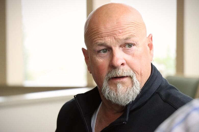 A man with a salt-and-pepper beard and moustache and a bald head speaks with someone else.