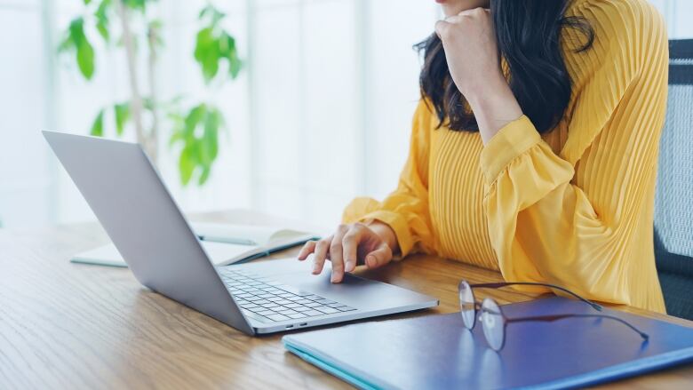 A woman in a yellow blouse is seen scrolling on her laptop.