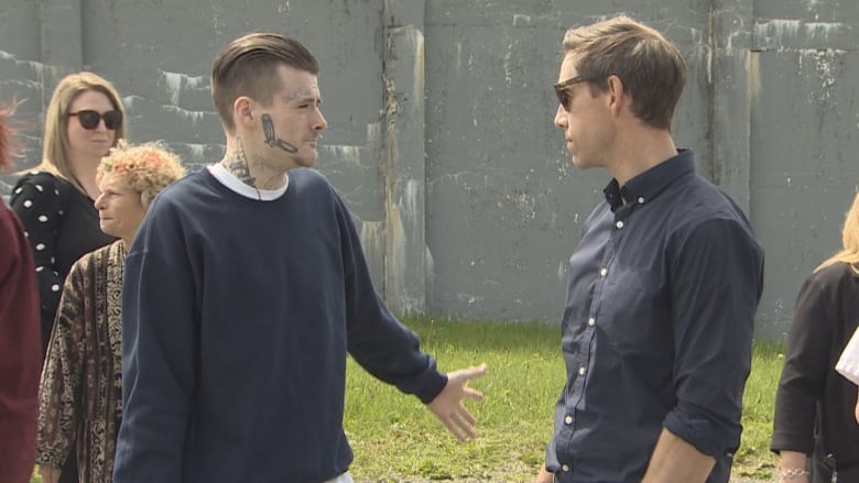 A man in prison garb shrugs as a tall man wearing a dress shirt and glasses looks on. His hands are in his pockets. 