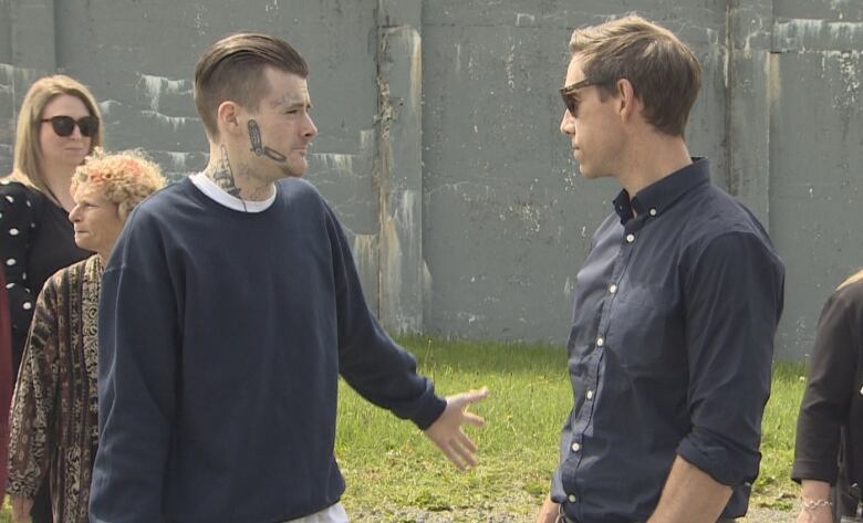 A man in prison garb shrugs as a tall man wearing a dress shirt and glasses looks on. His hands are in his pockets. 