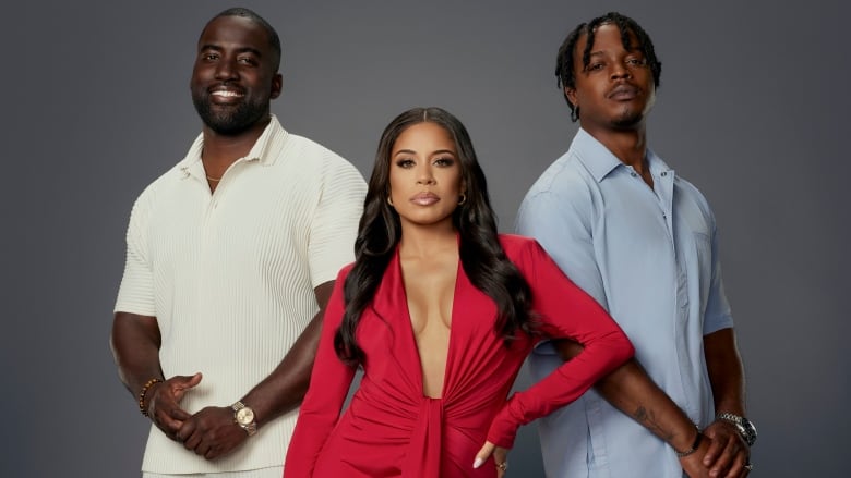 Two men and a woman pose in front of a grey background. The woman is in the centre, wearing a red dress. The man on the left wears white pants and a white shirt while the man on the right wears blue. 