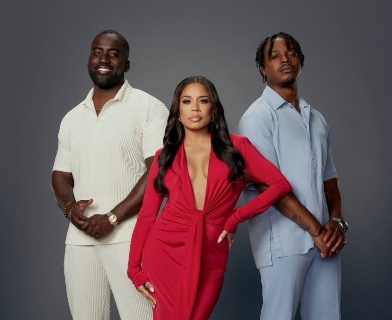 Two men and a woman pose in front of a grey background. The woman is in the centre, wearing a red dress. The man on the left wears white pants and a white shirt while the man on the right wears blue. 