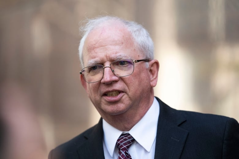 A white-haired man in a suit and tie is shown speaking in an outdoor photo.