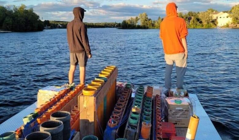 Fireworks sit on a dock waiting to be fired off while two people stare out a lake