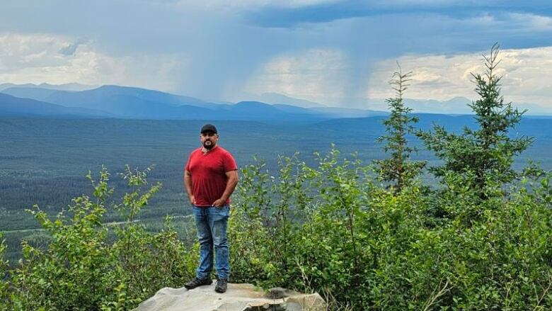 A man in a red shirt, blue jeans, and baseball cap, stands proudly on a rock above a forested valley with high hills and wisps of smoke in the background. 