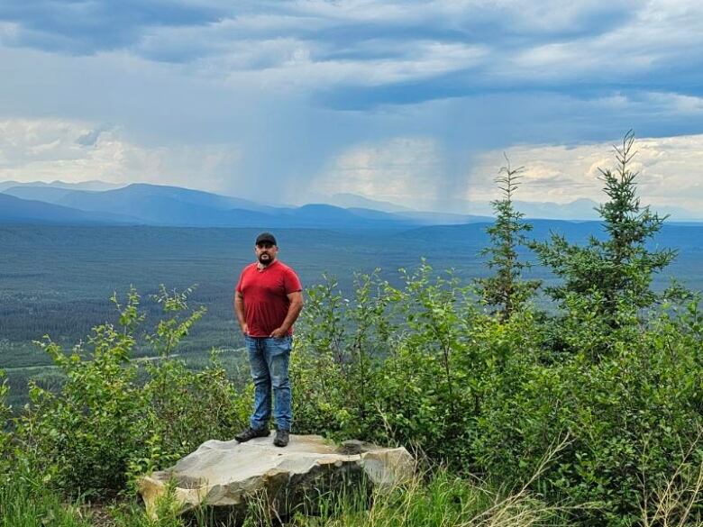 A man in a red shirt, blue jeans, and baseball cap, stands proudly on a rock above a forested valley with high hills and wisps of smoke in the background. 