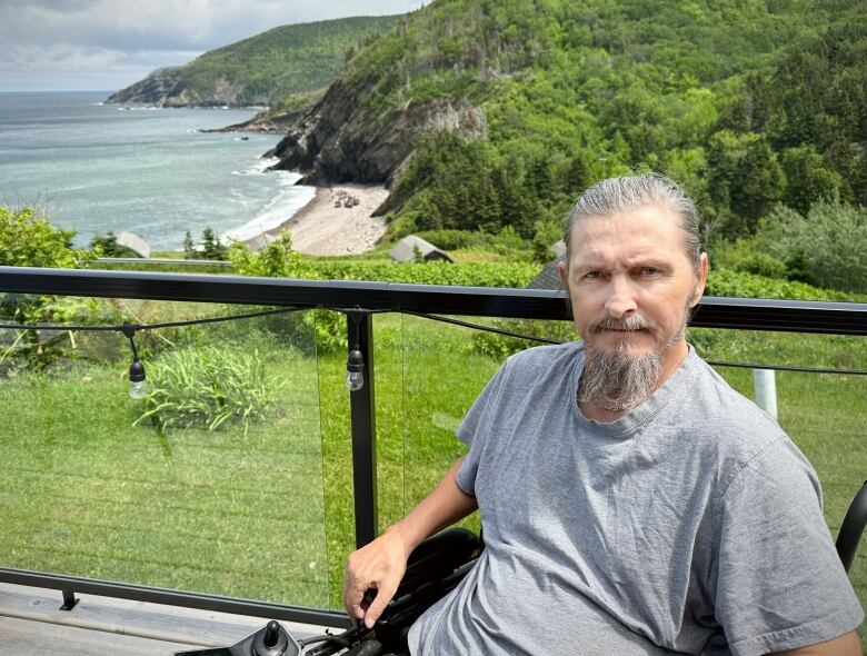 A man sits on a deck overlooking a beach and the ocean, with a mountain in the background.