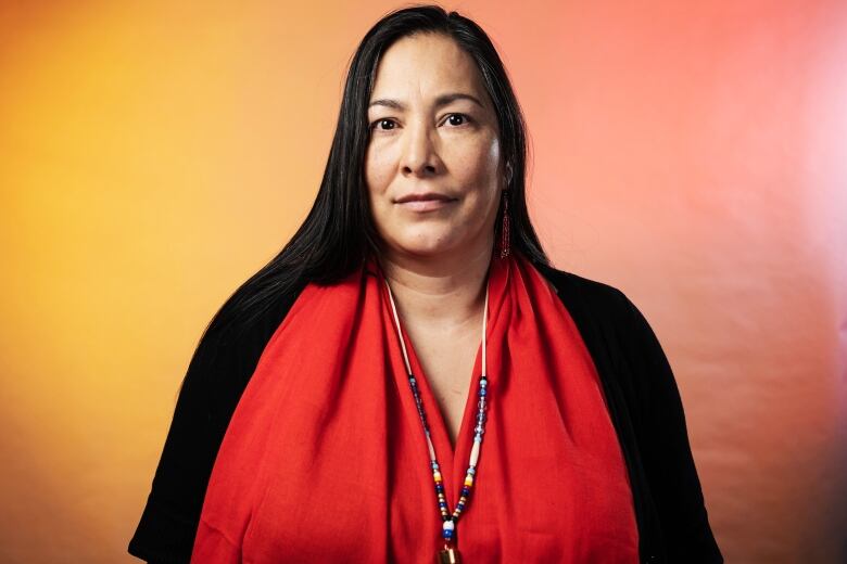 A woman with long black hair, wearing a black shirt and a bright red scarf, stares straight ahead into the camera.