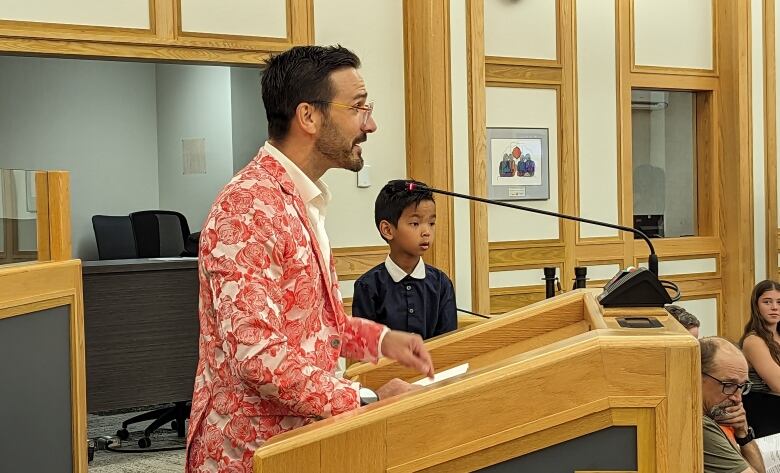 A man in a bright floral blazer speaks at a podium with a young boy sat beside him