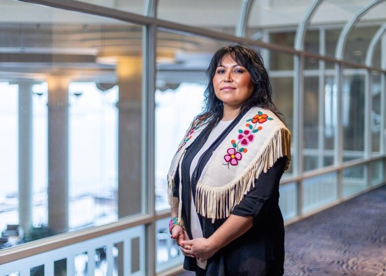 An Indigenous woman who is the chief of the Blueberry River First Nations stands in a dark dress and beaded white vest with flowered symbols on each shoulder in a bright room. 