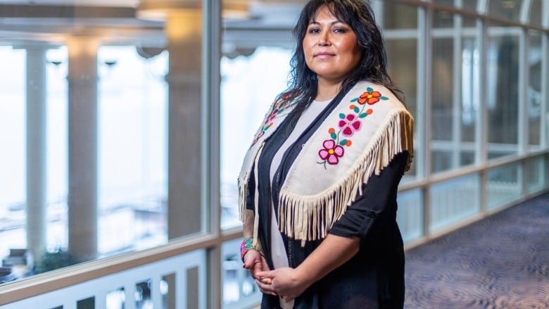 An Indigenous woman who is the chief of the Blueberry River First Nations stands in a dark dress and beaded white vest with flowered symbols on each shoulder in a bright room. 