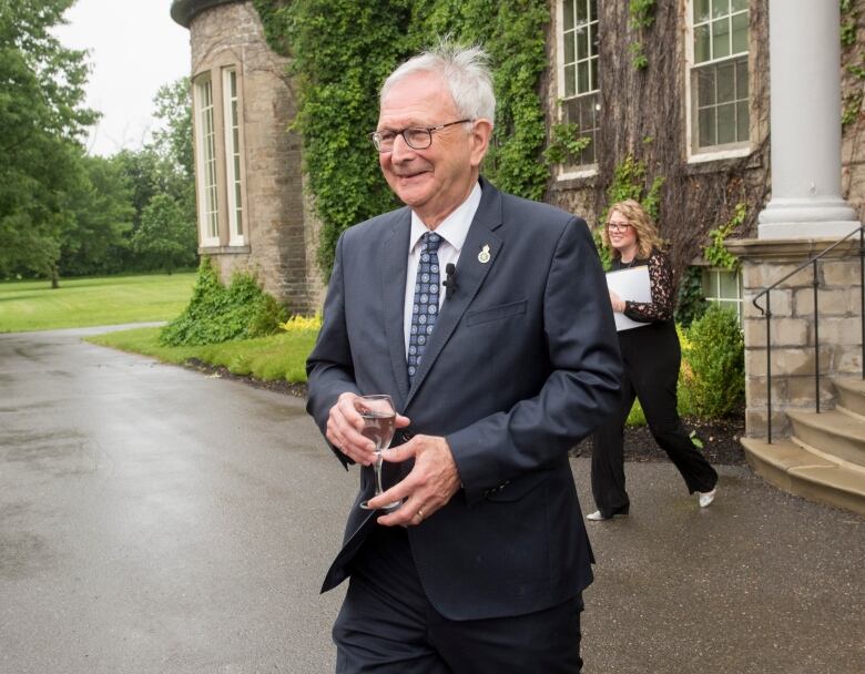 A smiling man in a suit holds a glass as he walks outside a stone building with concrete steps. 