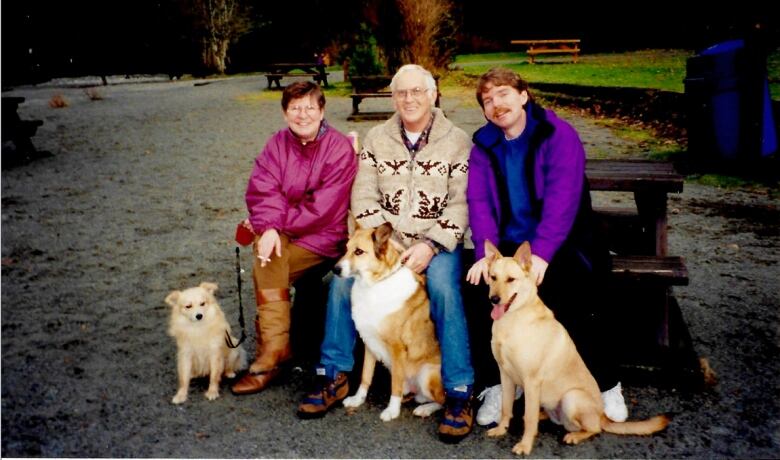 Three people sit on a park bench with their dogs.