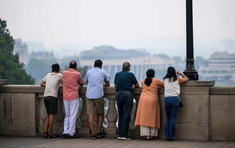 The backs of six people at a lookout over a river to another city. The air is smoky.