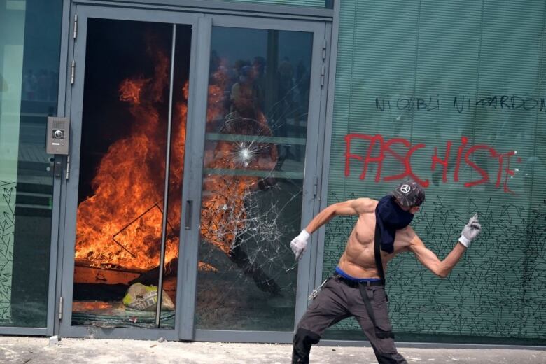 A man wearing a mask throws an object while a storefront burns in the background.