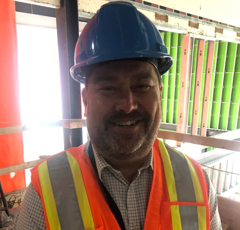 A man in his 40s is smiling at the camera. He is wearing a dress shirt and a hard hat. He is on a construction site with brick and an open wall behind him. 