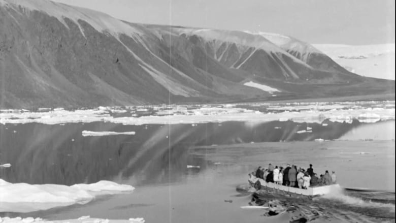 A black and white photo of a large open boat approaching a mountainous coast; the water is covered in ice.