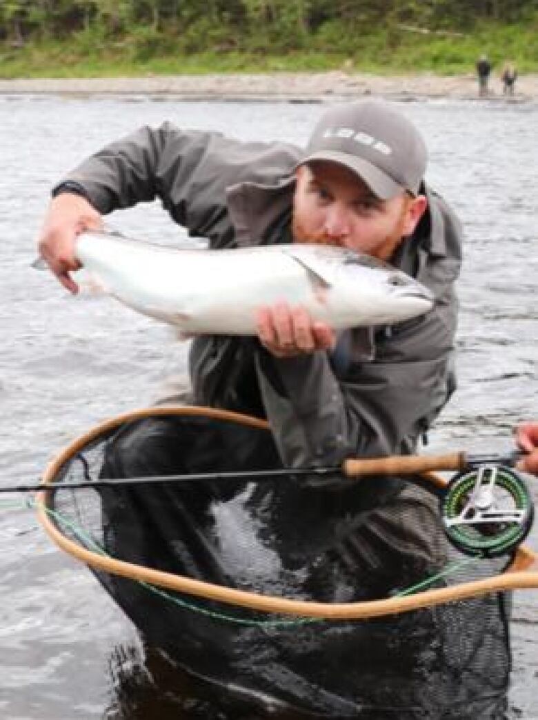 A man holding up a salmon while crouching in a river gives the fish a kiss on the top of its head.