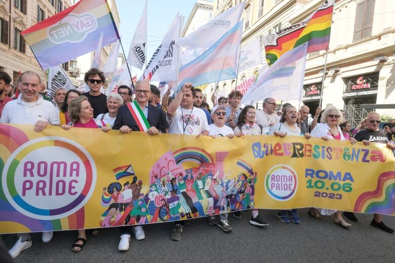 A man wearing a dark suit and a sash in the colours of the Italian flag smiles and stands among a group of cheering people behind a banner reading 'Roma Pride' at Rome's annual LGBTQ+ Pride parade. 