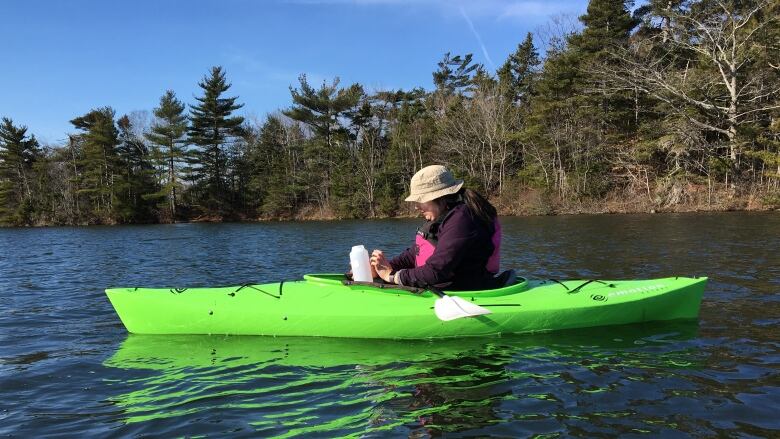 A person in a wide brim hat collects a water sample while in a kayak on a lake.