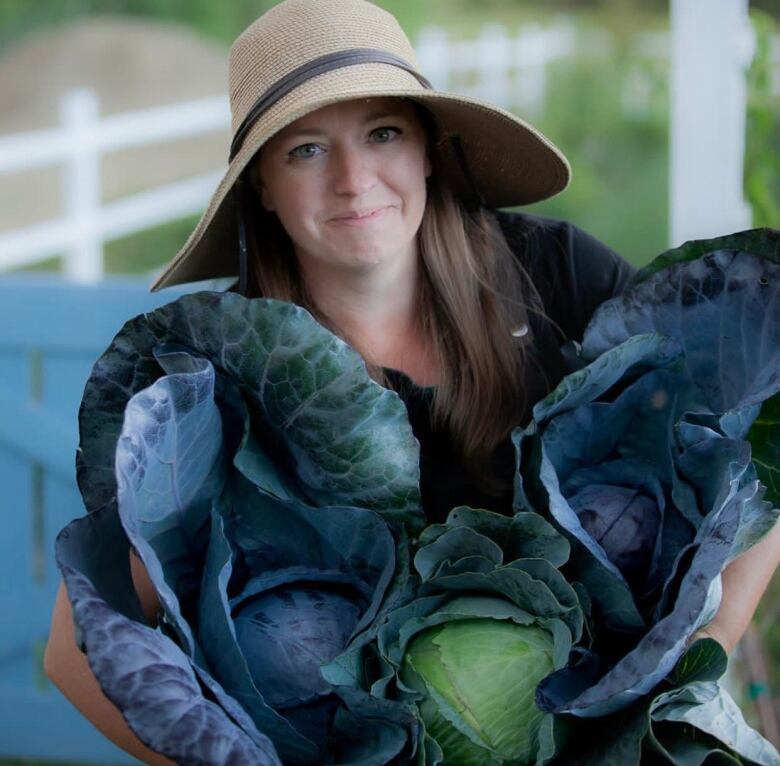 A woman in a wide brimmed hat holds two large cabbages. 