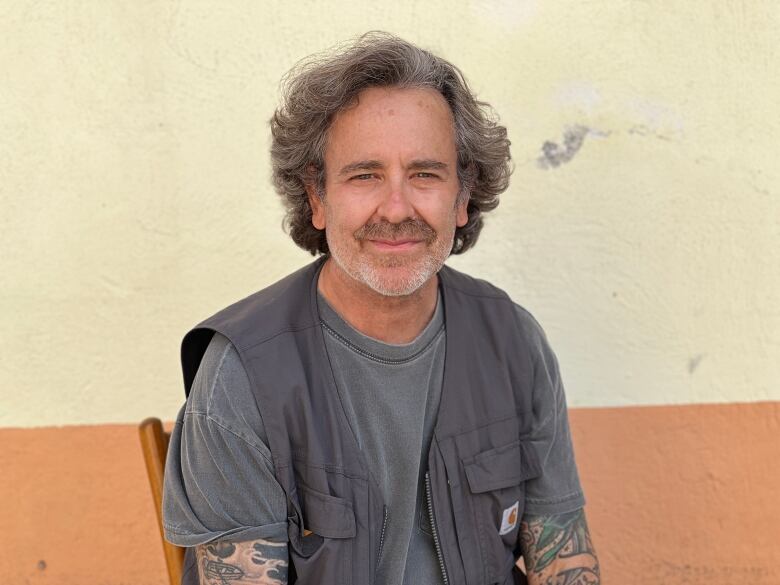 A man wearing a grey shirt and vest, with short, feathered hair, poses for a portrait as he sits in front of a yellow concrete wall.