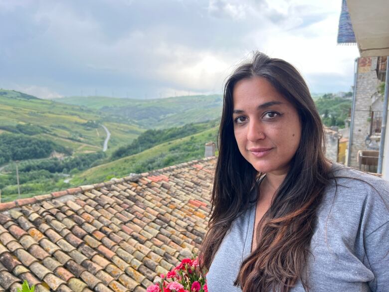 A woman with long dark hair smiles, as she looks into the camera, standing on a balcony with a terracotta roof and lush green hills in the background.