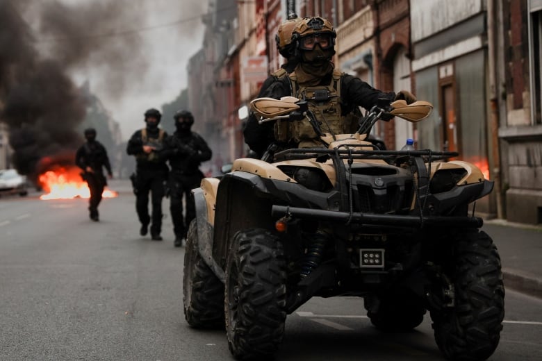 People in military gear ride an alternative vehicle in the street as other soldiers walk behind and a fire burns.