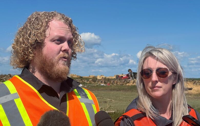 A man with curly hair stands next to a blonde woman. They are wearing safety vests. 