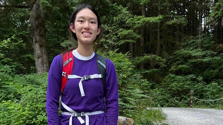 A teen girl in hiking clothes and a backpack smiles at the camera in a gravel parking lot with trees behind.