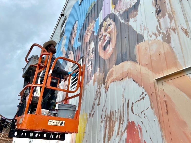 A man on a lift holding a paintbrush works on a large mural.