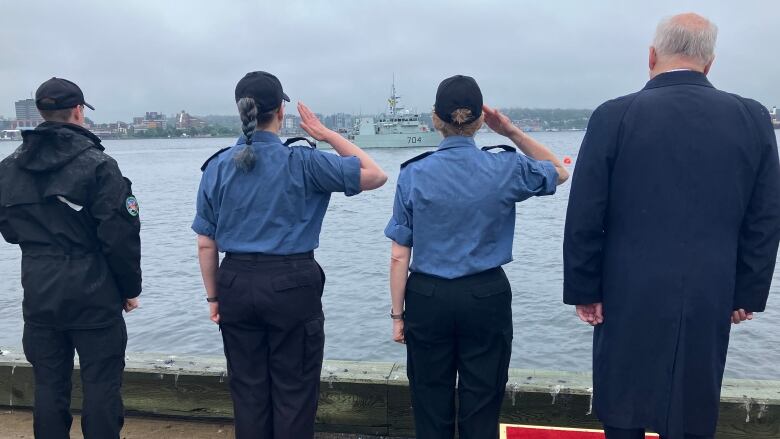 Three people in Navy uniform stand on the edge of the habour facing the water and saluting a ship in the distance. 