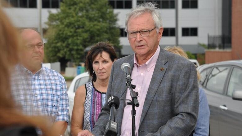 A man with grey hair speaking at a podium with others gathered behind him