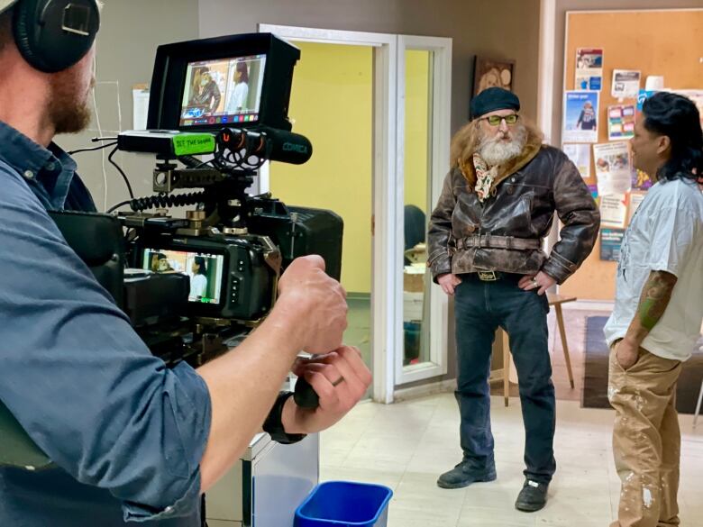 A man behind a video camera focuses on a man with a beard and red hair and a second man with dark hair.