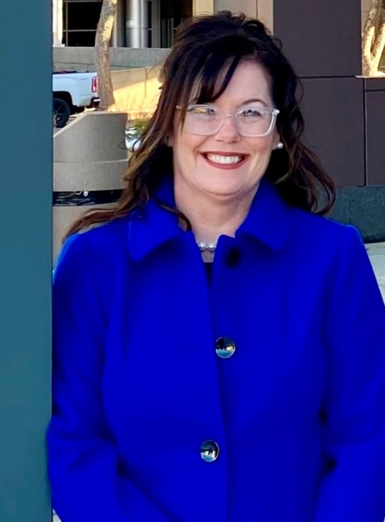 A woman wearing glasses stands and smiles wearing a blue peacoat.