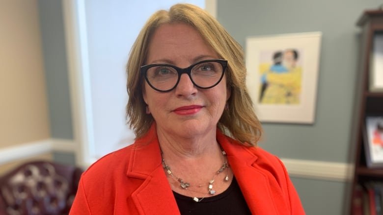 A smiling woman with glasses wearing a red coat stands in an office space.