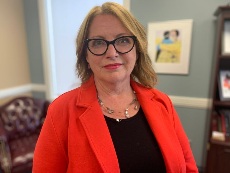 A smiling woman with glasses wearing a red coat stands in an office space.